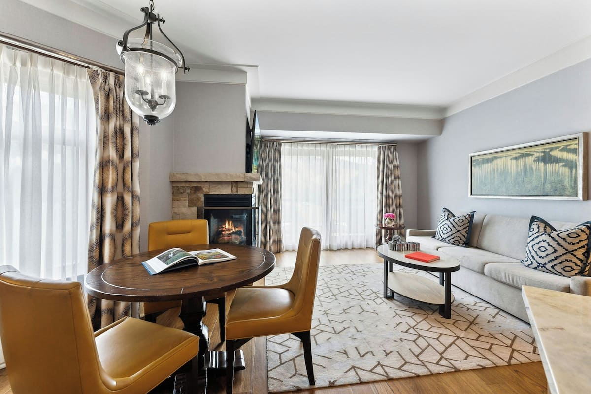 A modern living room with a fireplace, a round dining table with chairs, a sofa with patterned cushions, and a large painting on the wall.