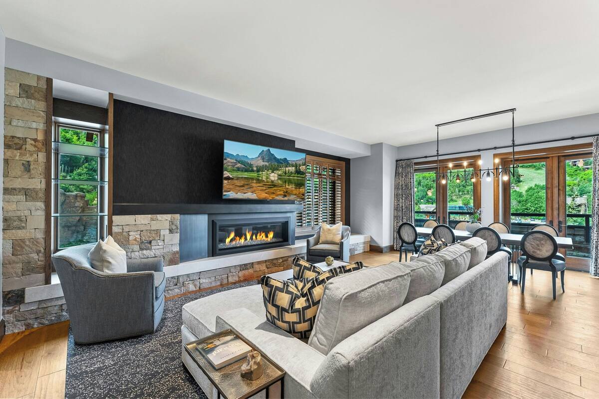 A modern living room with a fireplace, stone walls, and large windows offering a view of greenery and mountains.