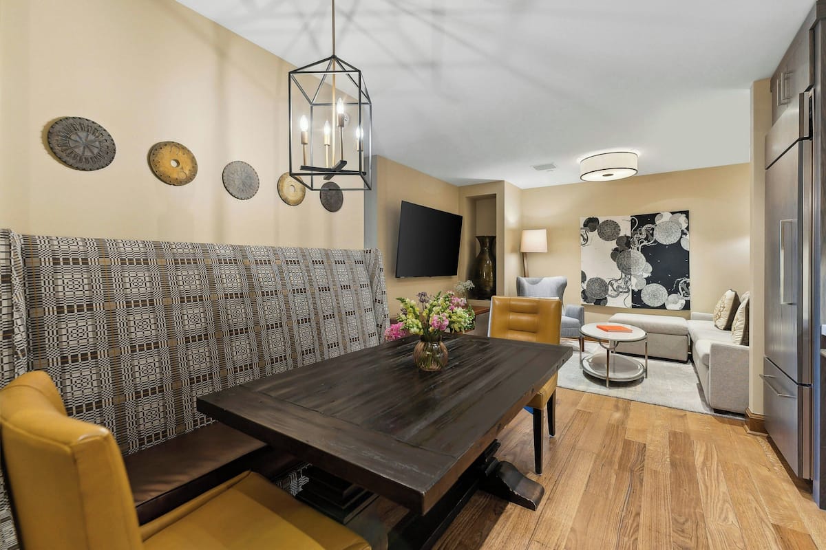 A modern living room with a dark wooden dining table, a sofa, and a TV mounted on the wall.