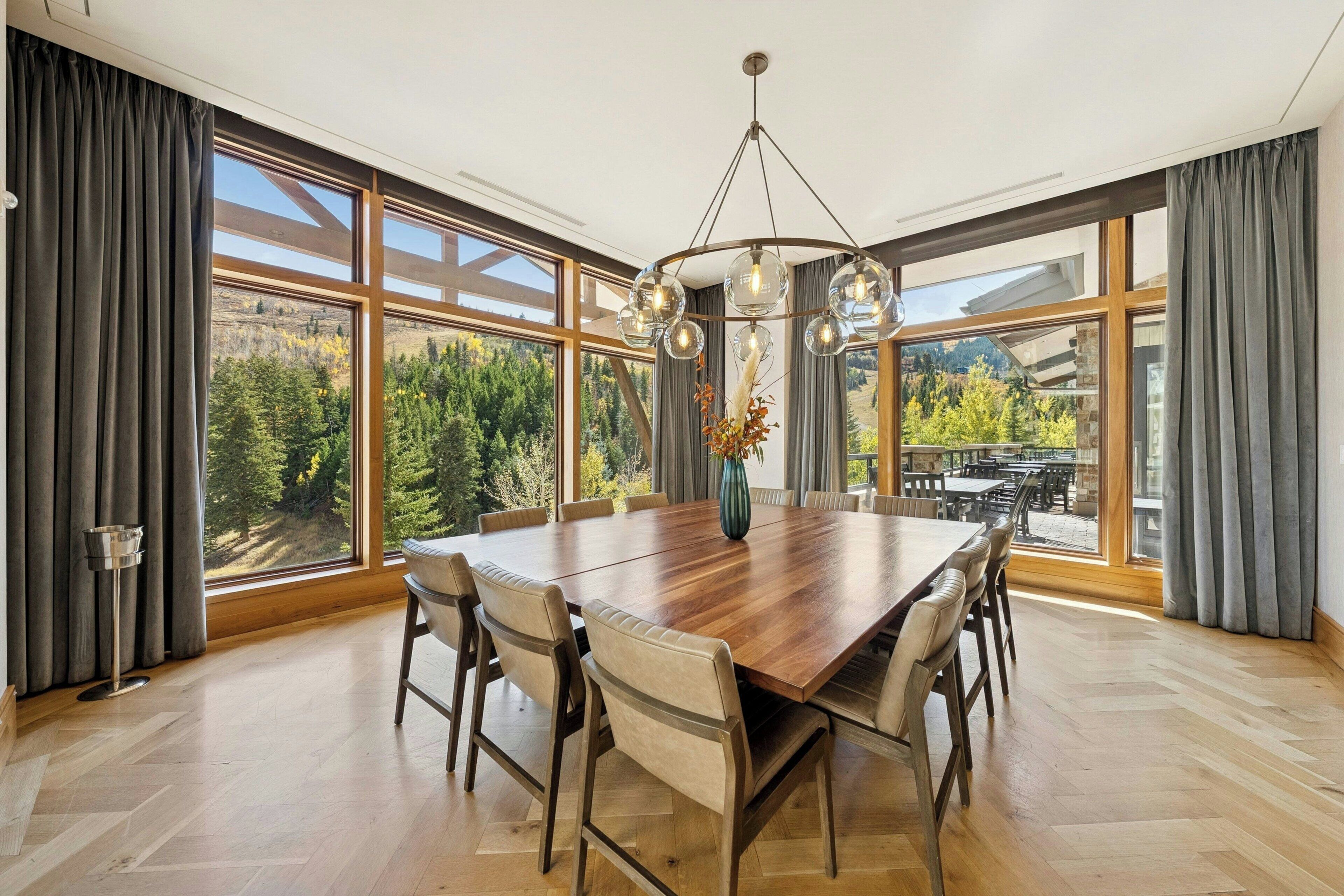 A dining room with a large wooden table, six chairs, a vase with flowers, and large windows with curtains.
