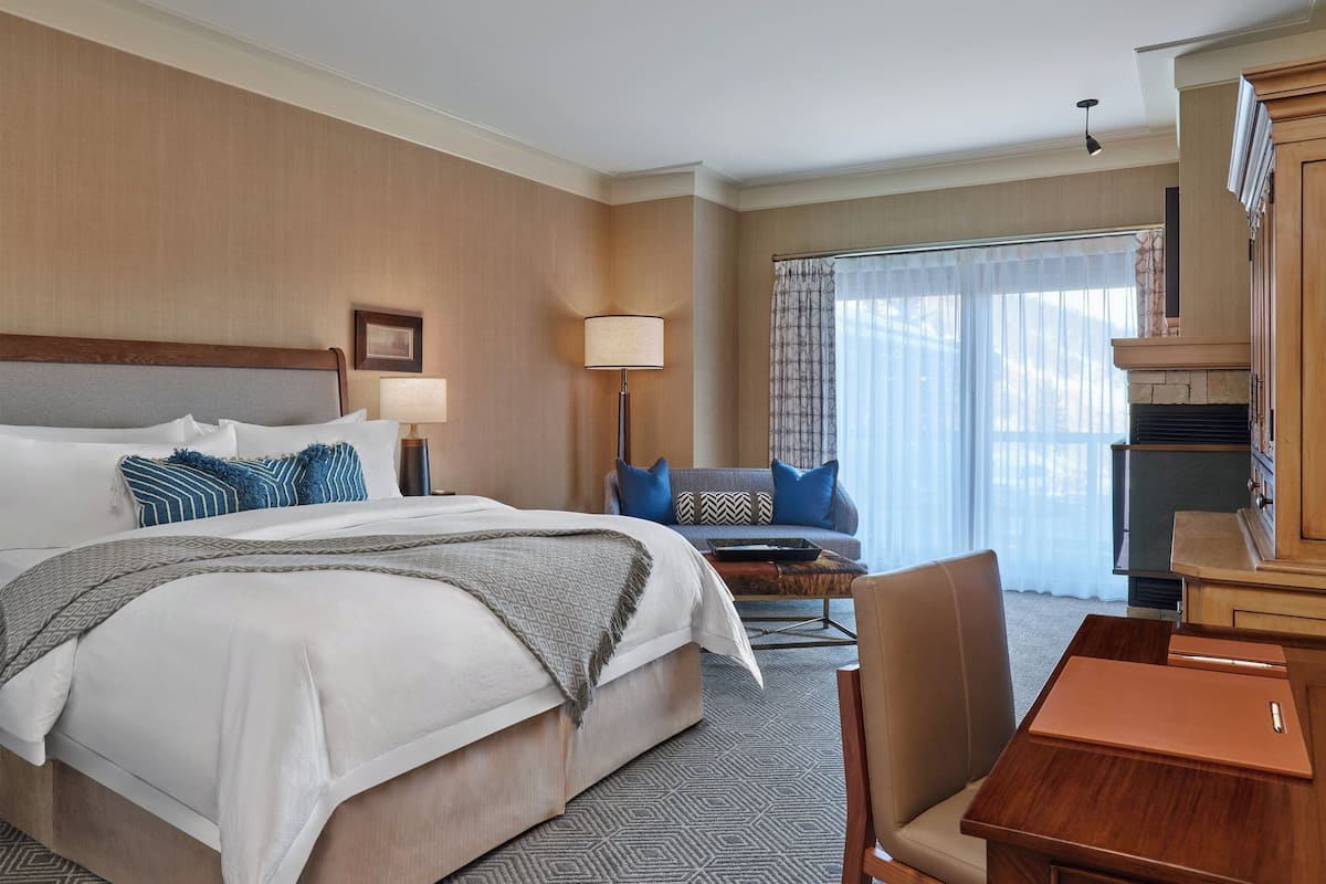 A hotel room with a large bed, a desk, a chair, a lamp, and a fireplace.