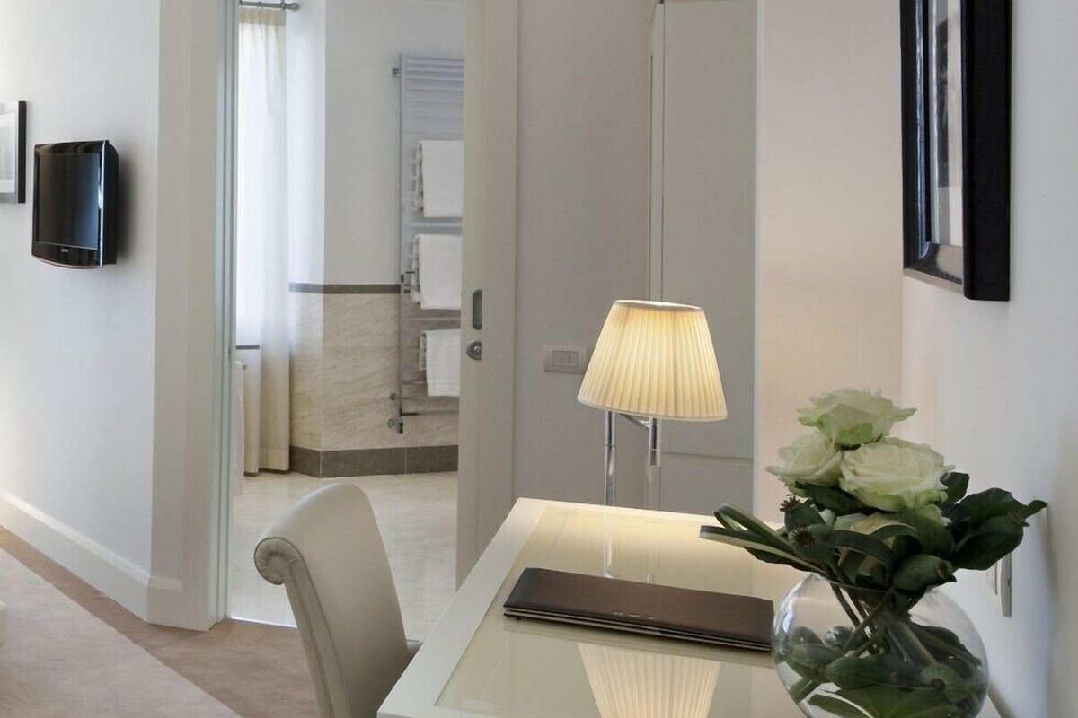 A hotel room with a glass table, a lamp, a vase with flowers, a television, and a door leading to a bathroom.