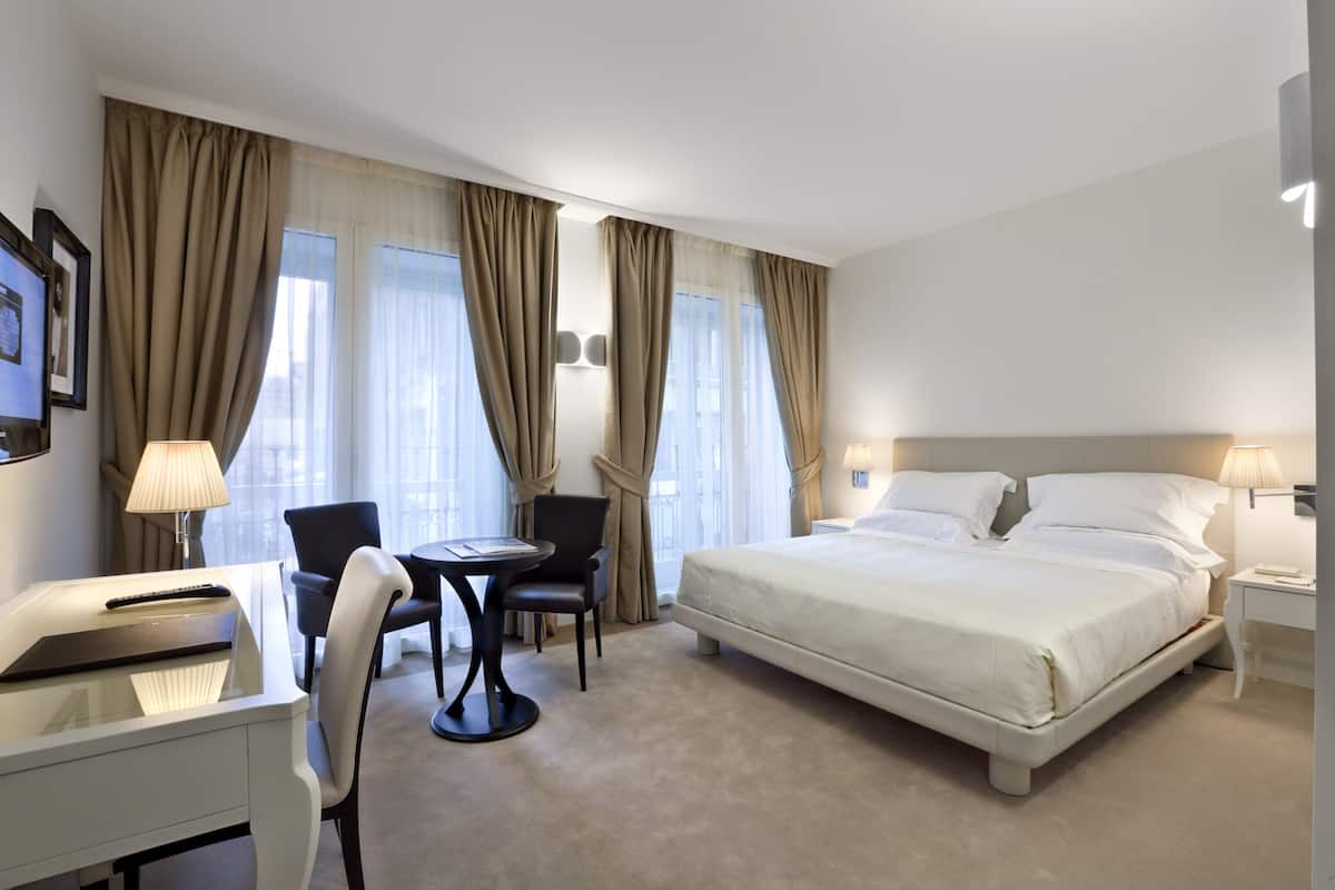 Une chambre d’hôtel avec un lit, un bureau, une chaise et deux fenêtres équipées de rideaux.