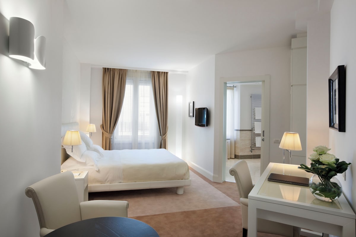 A modern hotel room with a bed, a desk, a chair, and a table. There is a TV mounted on the wall and a vase with flowers on the table.