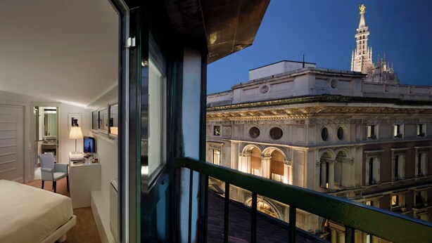 Habitación de hotel con cama, escritorio, silla y balcón con vista a un edificio histórico.