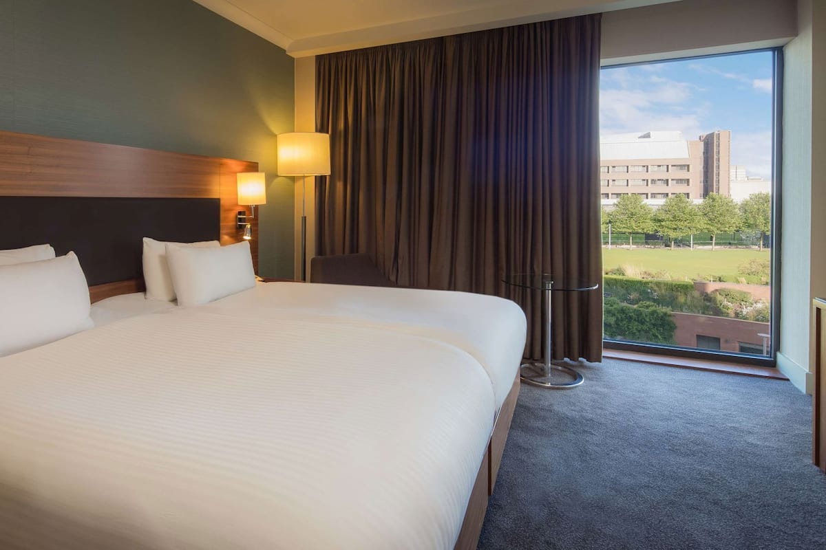 A hotel room with a large bed, a desk, a chair, a lamp, and a view of the city.
