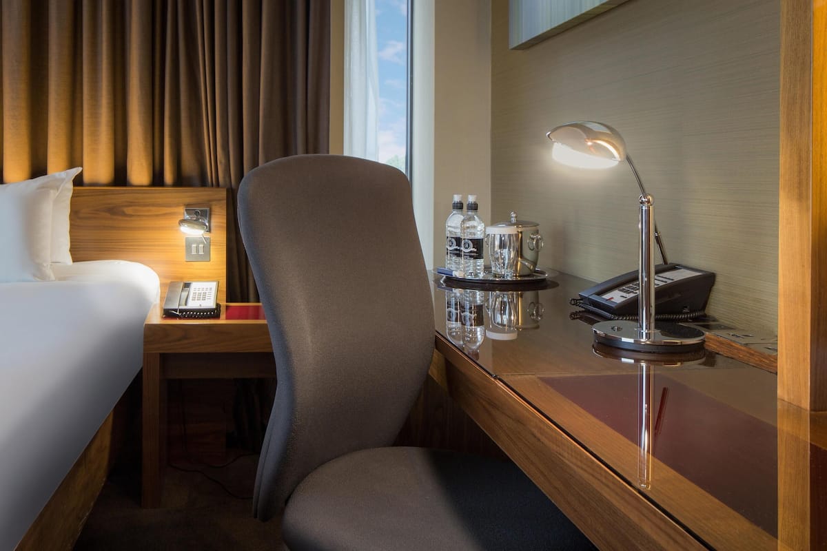 A hotel room with a bed, a desk with a lamp, a chair, and a minibar.