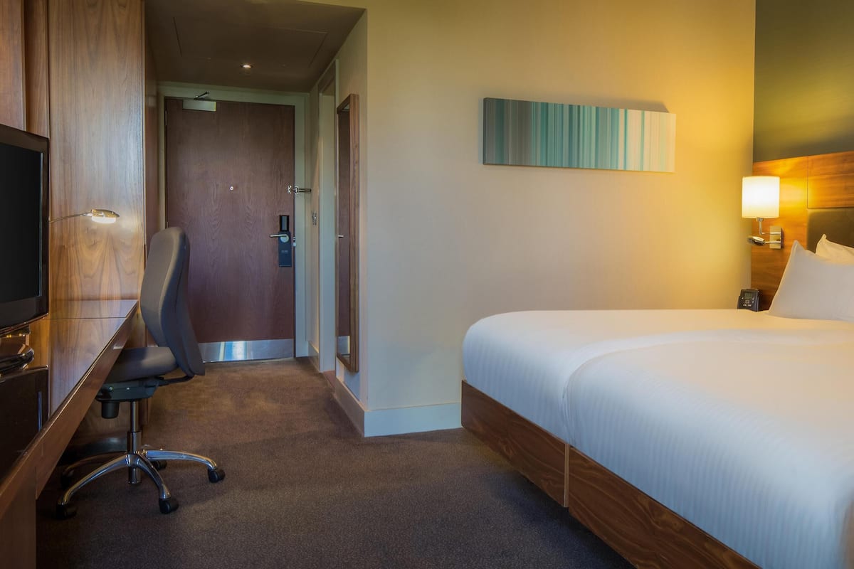 A hotel room with a bed, a desk with a chair, a television, and a painting on the wall.