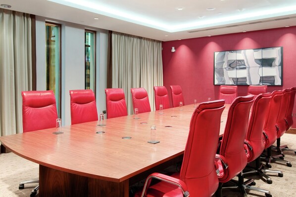 Une salle de conférence avec une longue table en bois et des chaises rouges, de grandes fenêtres avec des rideaux, et un mur d’accent rouge orné d’un tableau.