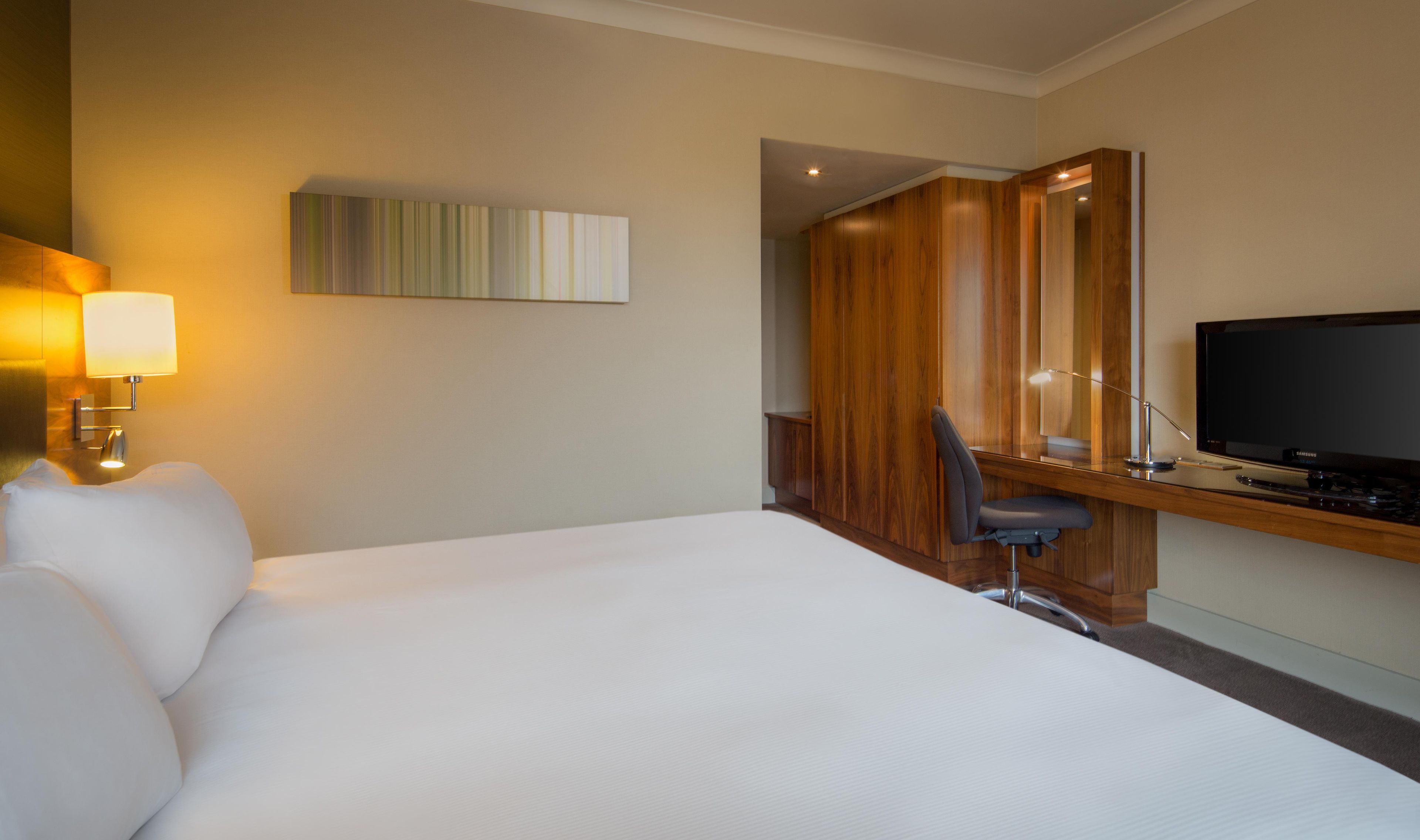 A hotel room with a large bed, a desk with a chair, a television, and a wall-mounted lamp.