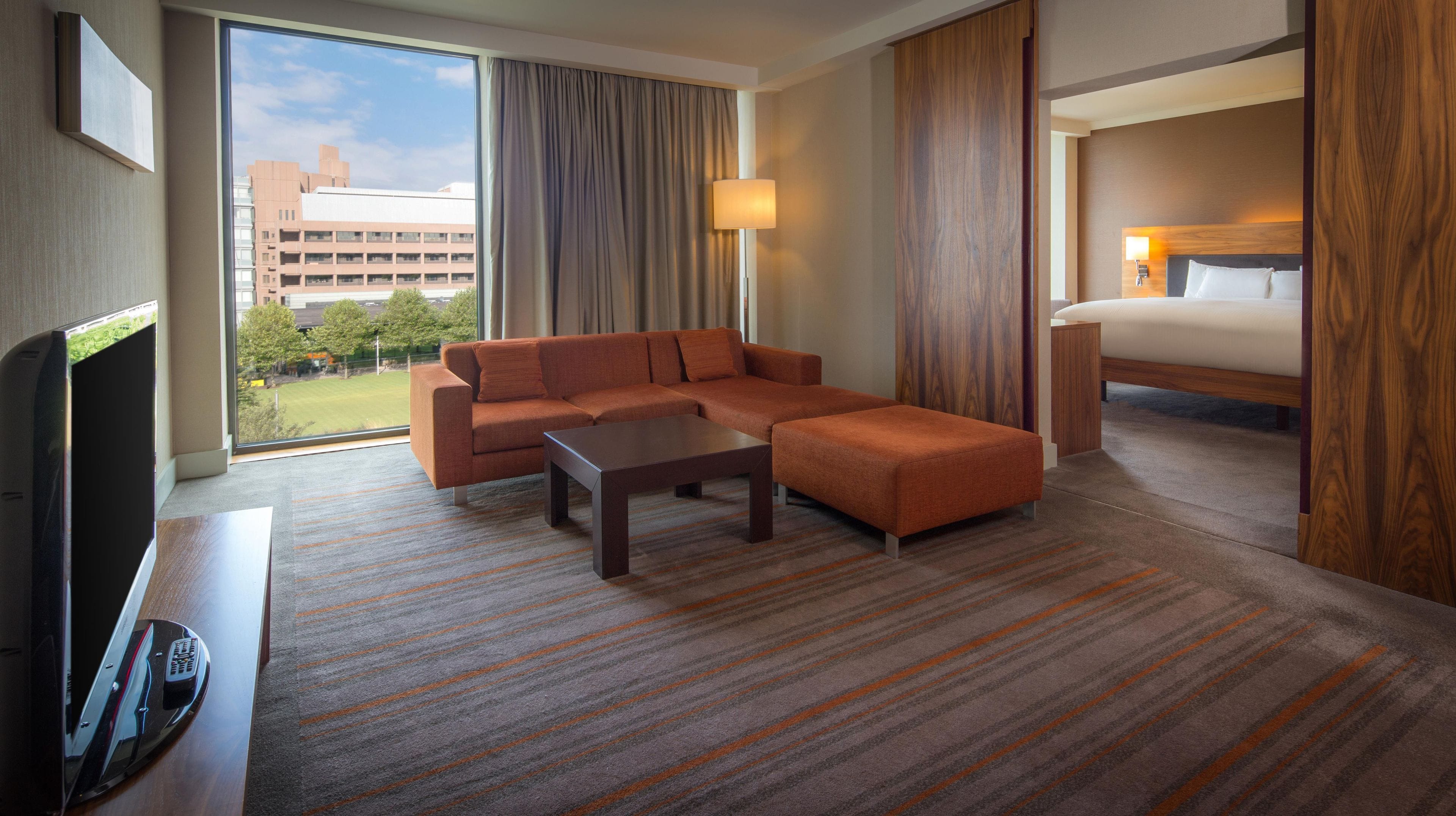 Habitación de hotel con sofá modular, cama individual, televisor de pantalla plana y ventana grande con vistas a edificios y árboles.