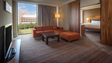 Habitación de hotel con sofá modular, cama individual, televisor de pantalla plana y ventana grande con vistas a edificios y árboles.