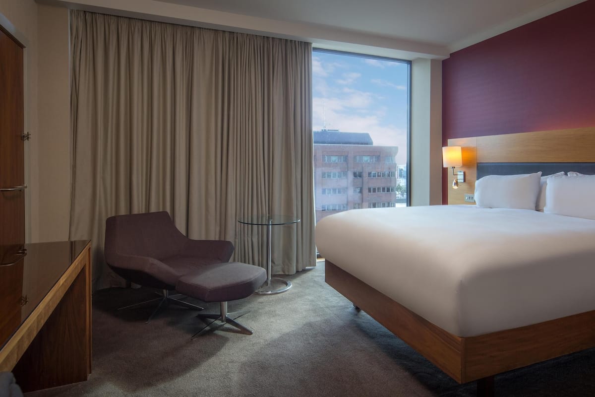 Une chambre d’hôtel avec un grand lit, un bureau avec une chaise, une télévision, une fenêtre avec des rideaux et une vue sur les bâtiments environnants.