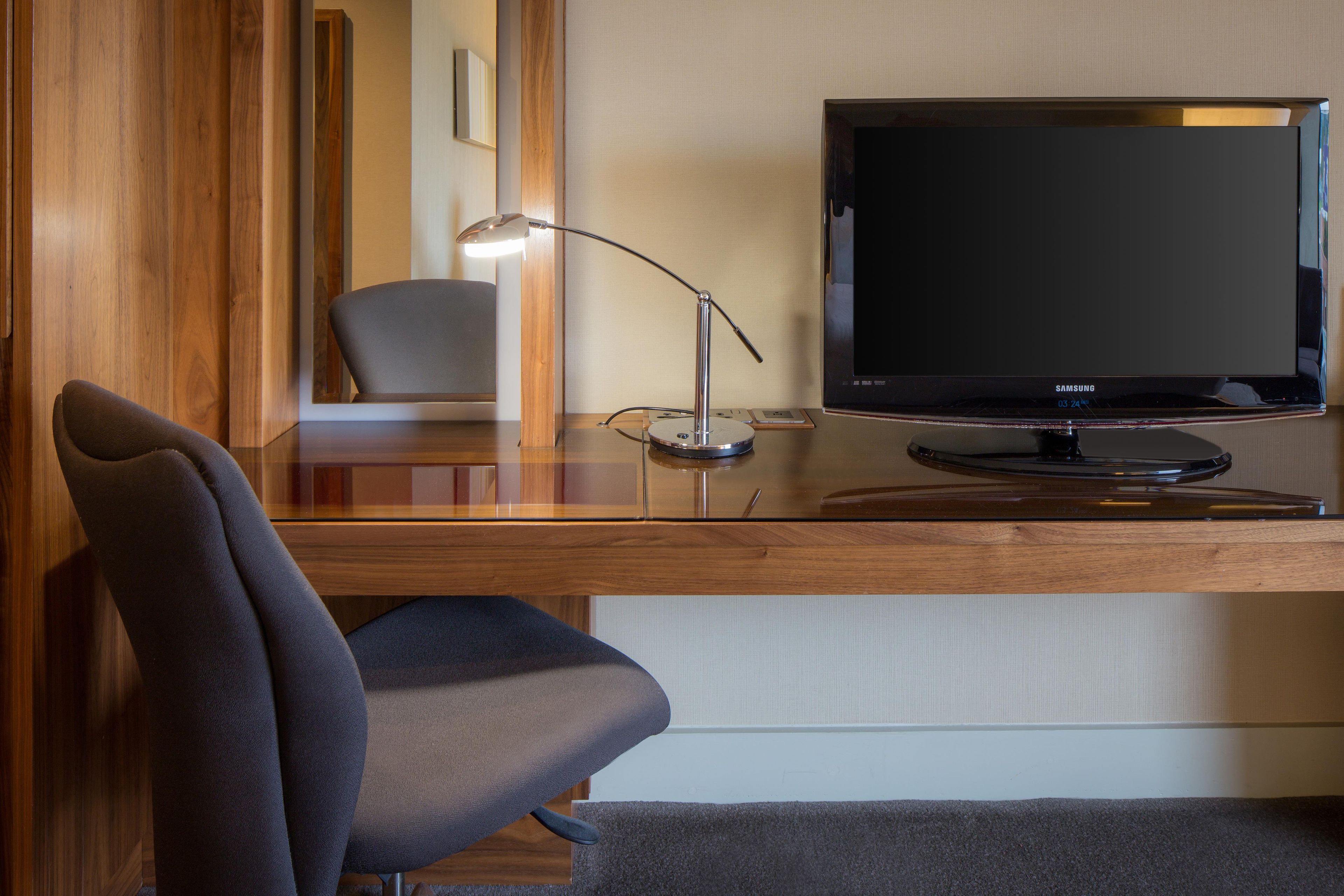 A hotel room with a wooden desk, a modern chair, a flat-screen TV, and a desk lamp.