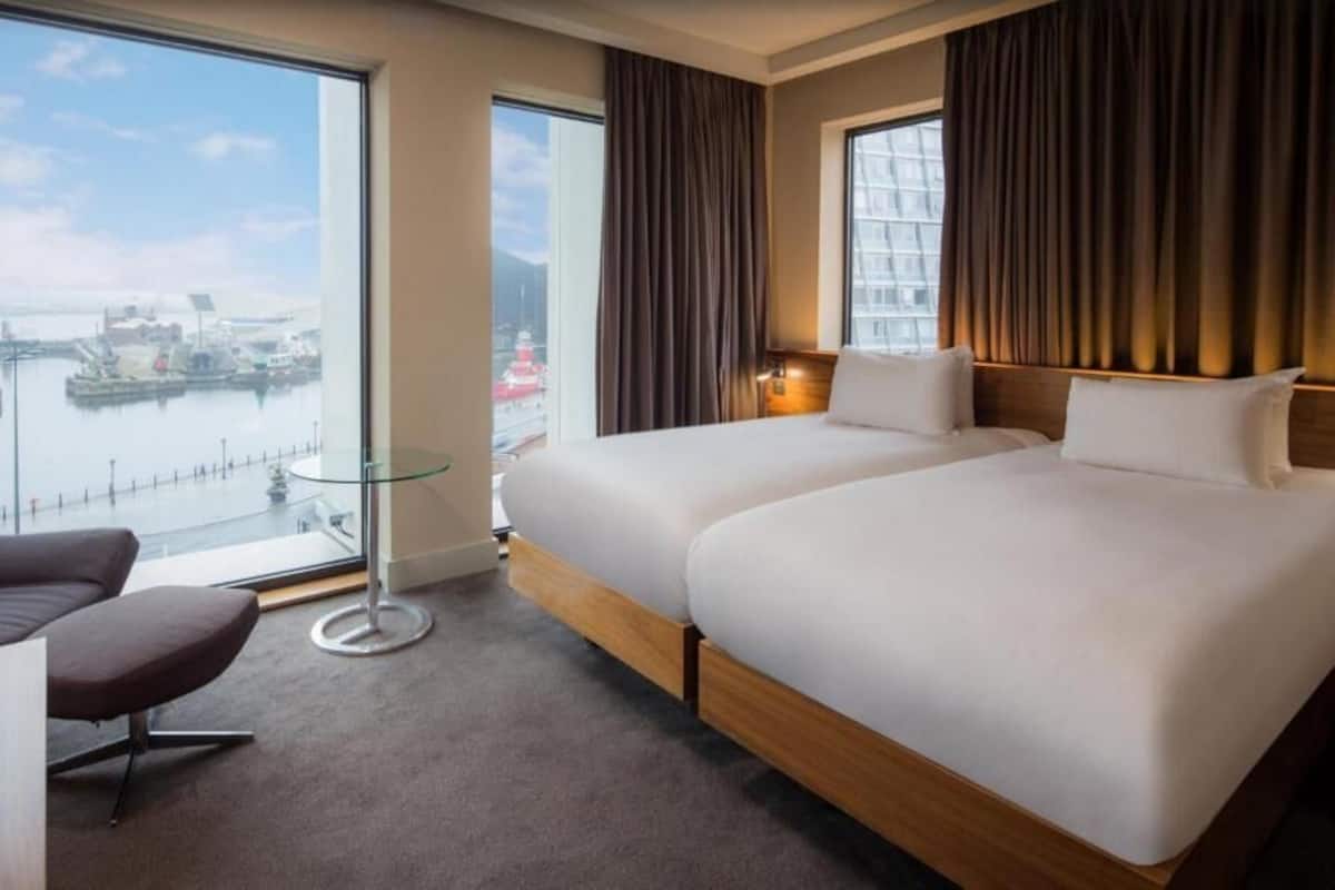 A hotel room with two beds, a desk, a chair, and a large window with a view of a harbor.