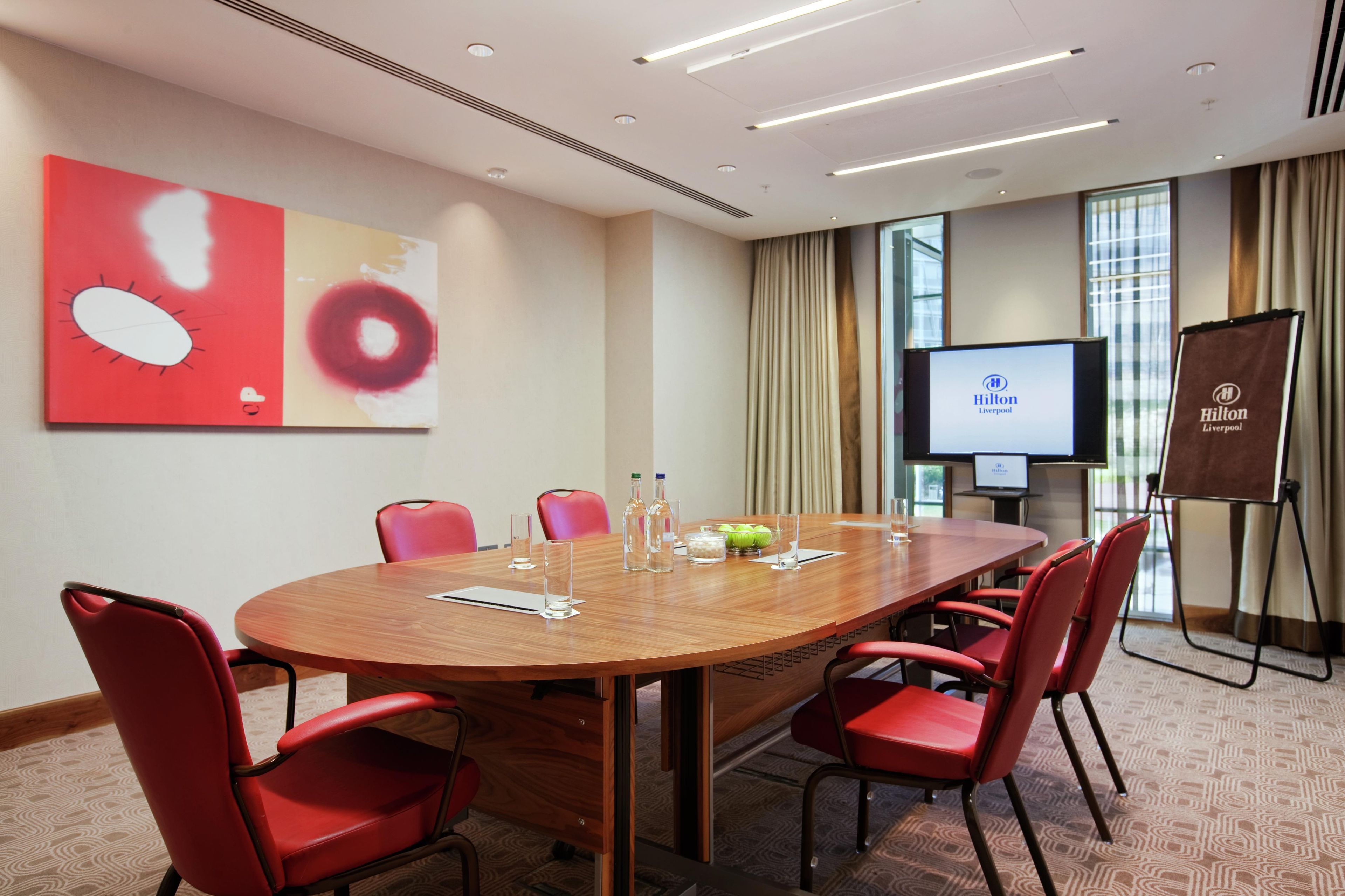 Une salle de conférence avec une table en bois, des chaises rouges, un écran de projection et une banderole à l’effigie de l’hôtel Hilton de Liverpool.