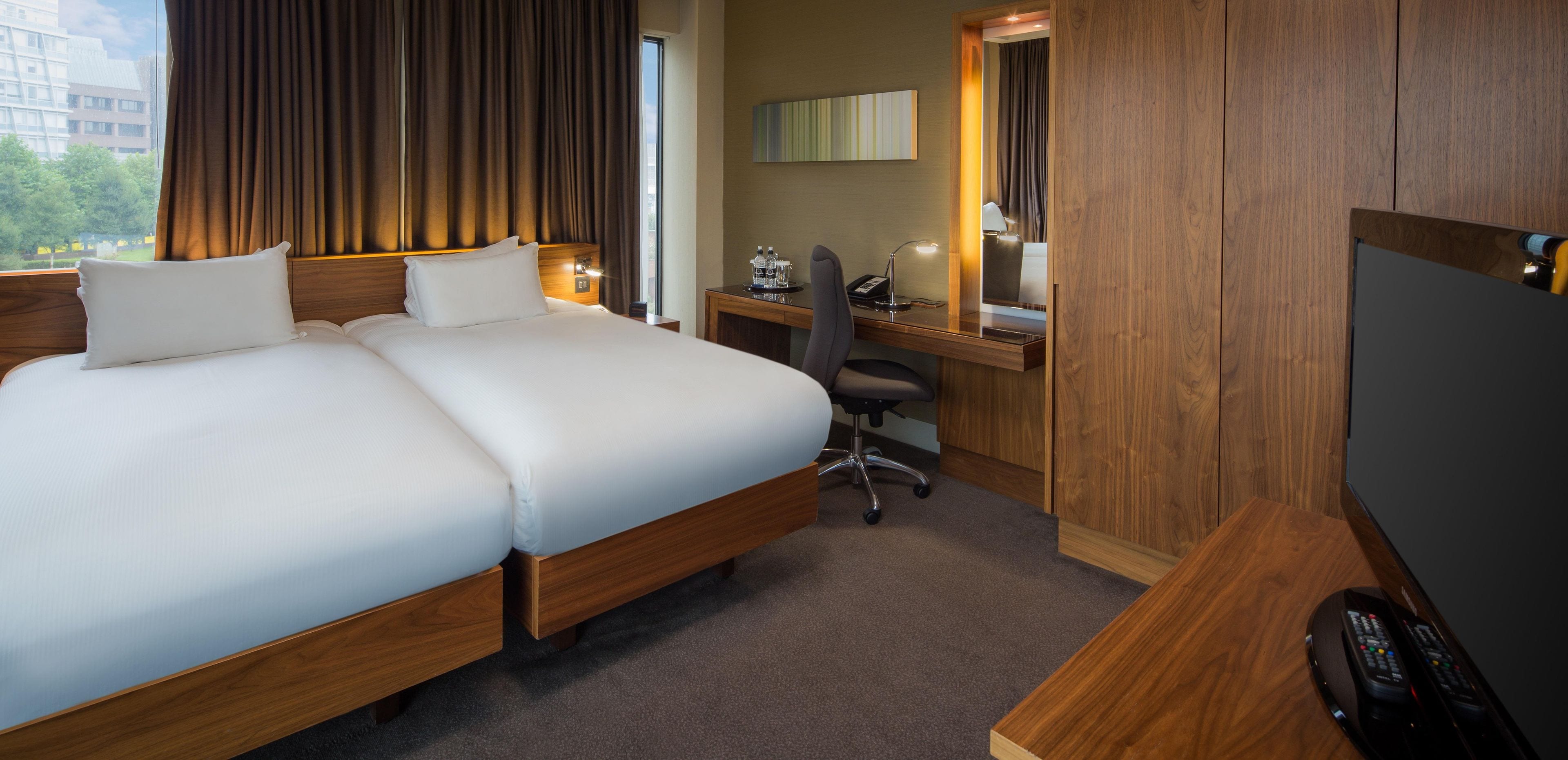 A hotel room with a large bed, a desk with a computer, a chair, a TV, and a window with curtains.