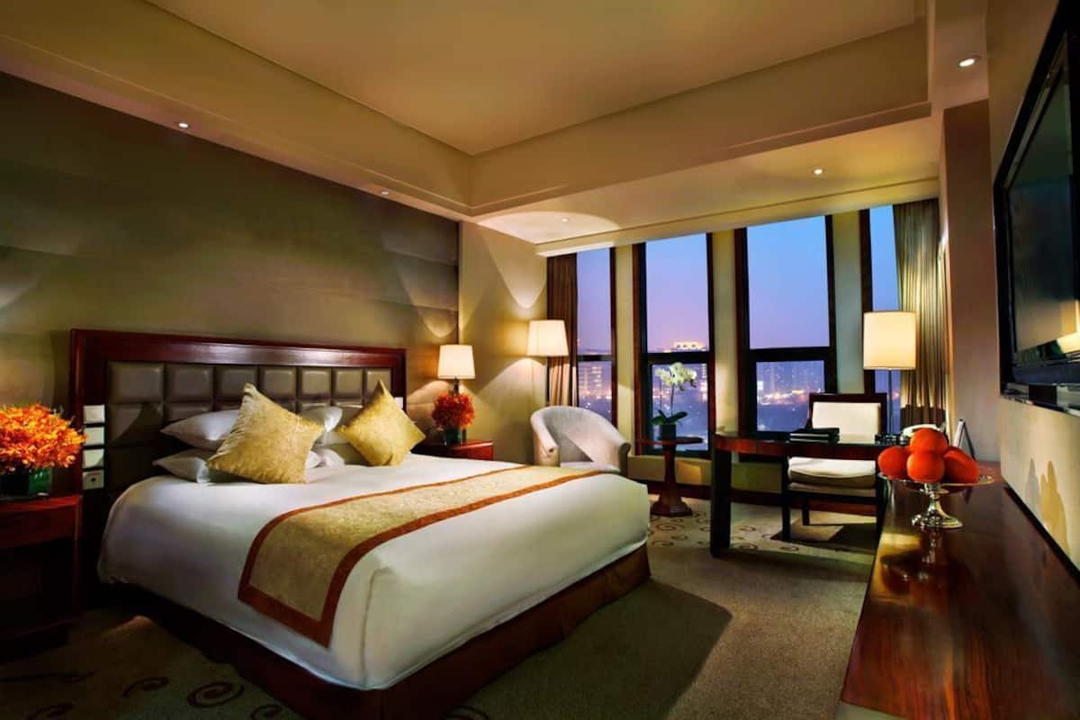 A hotel room with a large bed, two bedside tables, a desk, a chair, and a large window with a city view.