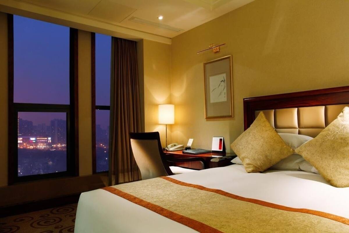 A hotel room with a large bed, a desk with a lamp, a phone, and a view of the cityscape at dusk.