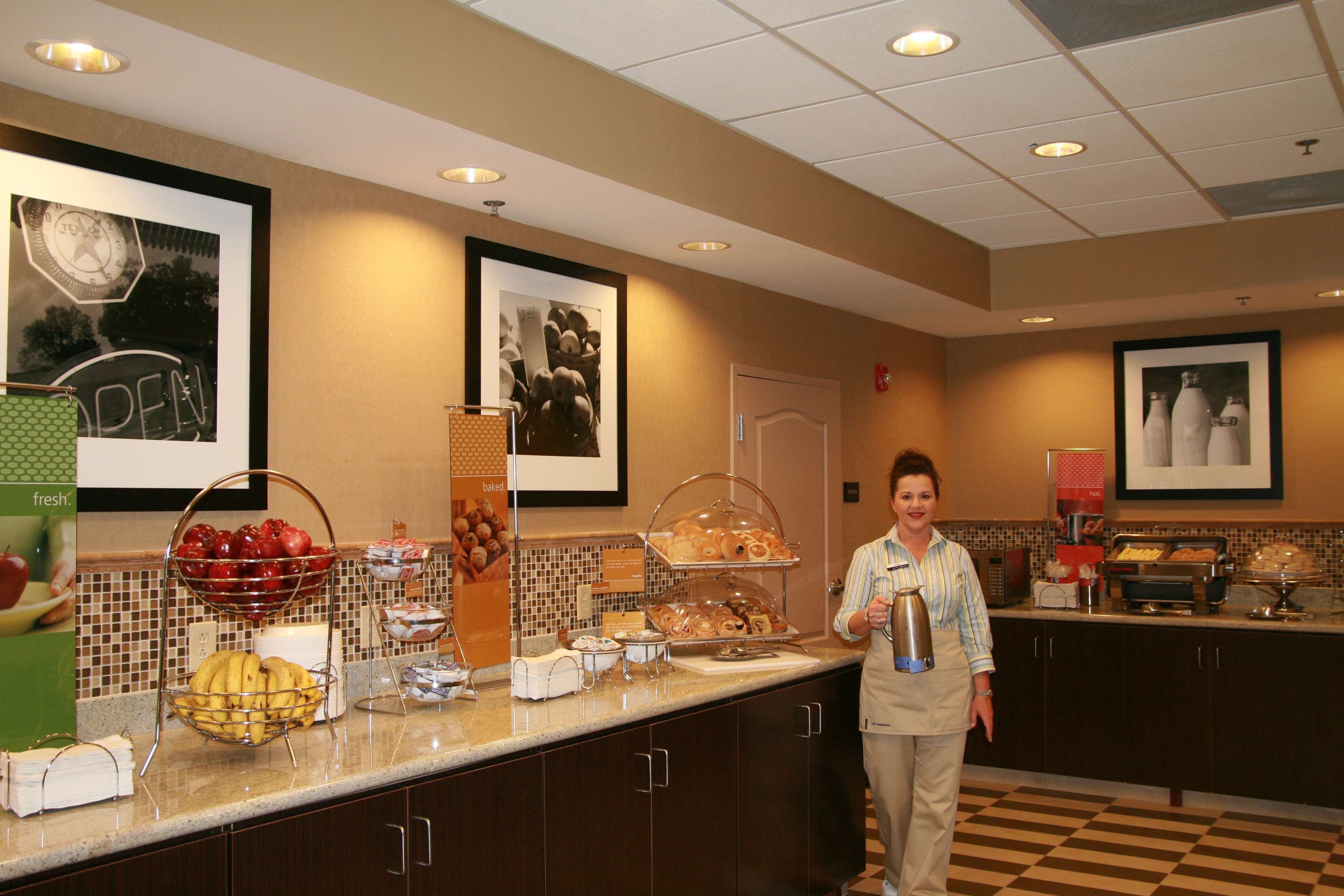 A buffet counter with various food items, a person standing, and framed pictures on the wall.