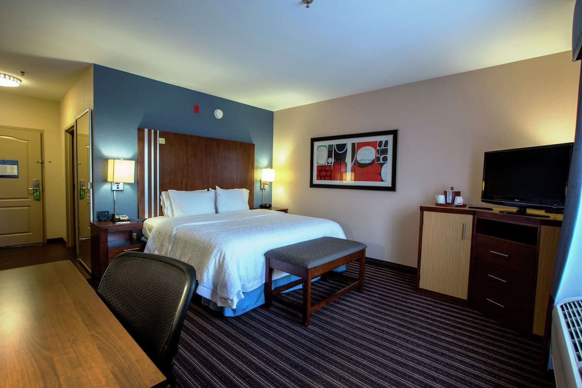 A hotel room with a bed, bedside table, lamp, TV, desk, chair, and a framed picture on the wall.