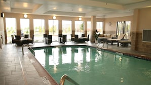 An indoor swimming pool with a tiled floor, a pool ladder, and several lounge chairs and tables around the pool area.