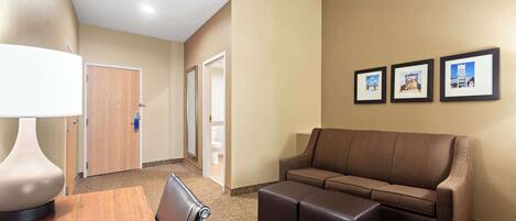 A hotel room with a brown sofa, a wooden table, and a lamp. There are three framed pictures on the wall.