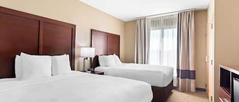 Une chambre d’hôtel avec deux lits, une tête de lit en bois, une table de chevet avec une lampe, un téléviseur et une fenêtre avec des rideaux.