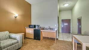 A compact kitchenette with a microwave, sink, and cabinets, a small sofa, and a dining table in a room with patterned carpet and a brown accent wall.