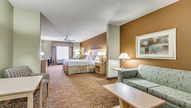 A hotel room with a bed, a sofa, a desk, and a chair. There is a ceiling fan, a window with curtains, and a framed picture on the wall. - Holiday Inn Express Hotel Suites Lubbock Southwest-Wolfforth by IHG (Wolfforth)