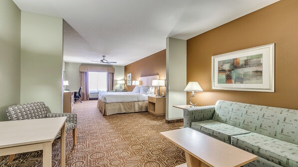 A hotel room with a bed, a sofa, a desk, and a chair. There is a ceiling fan, a window with curtains, and a framed picture on the wall.
