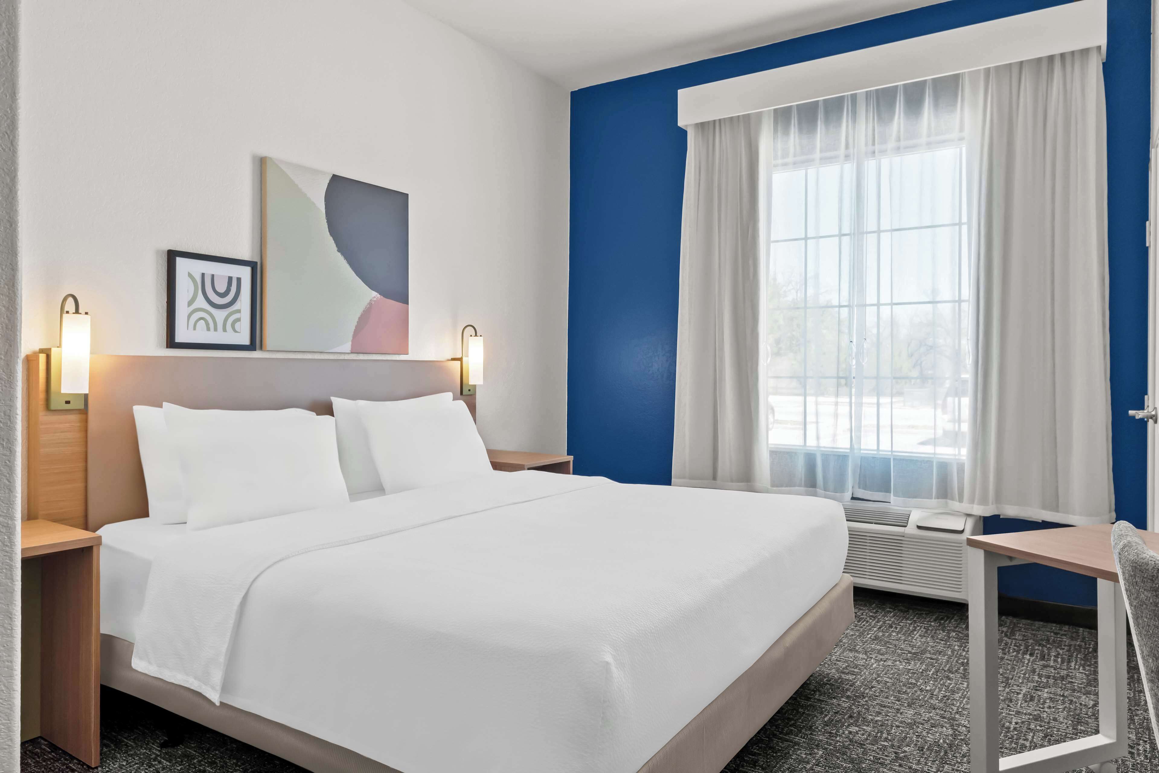 A hotel room with a large bed, a desk, and a chair. There is a window with curtains and a painting on the wall.
