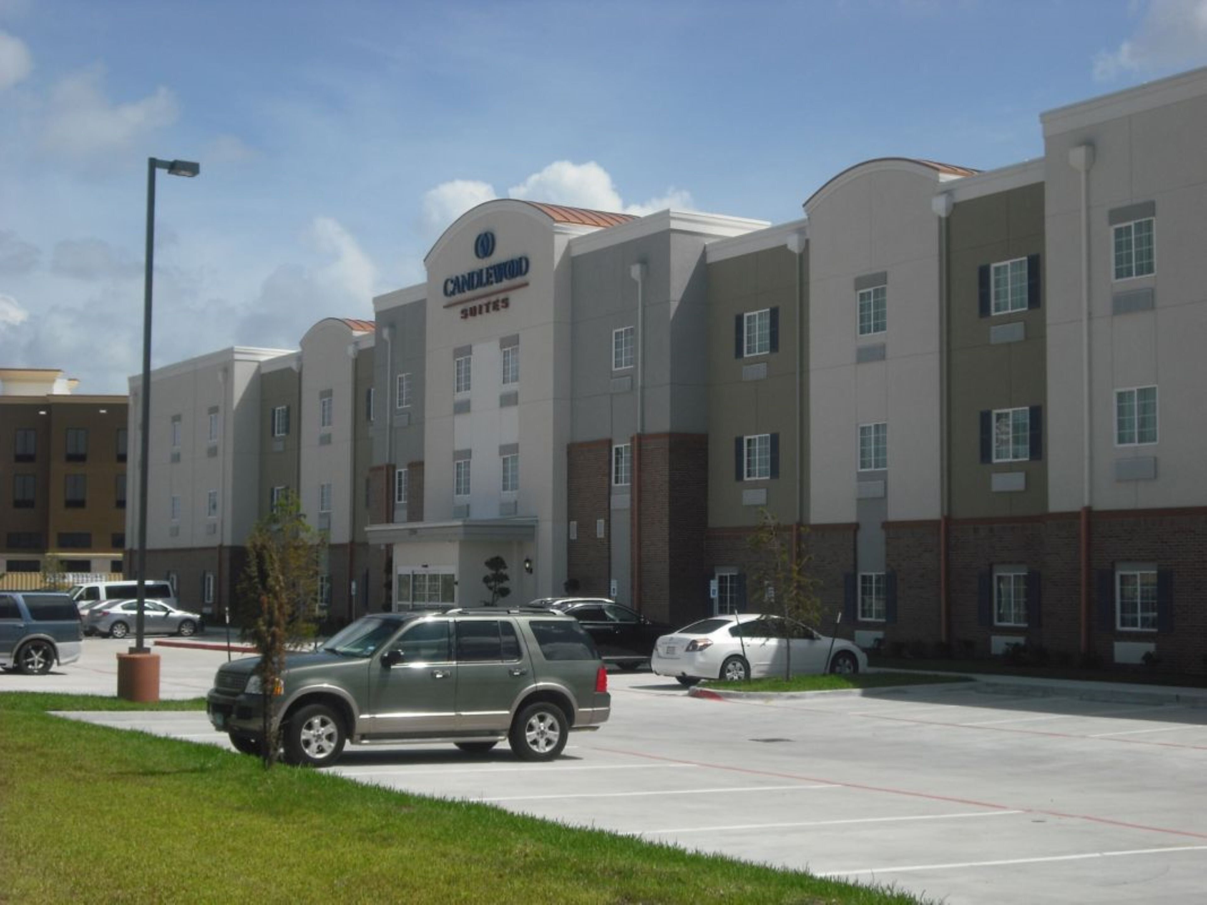 A multi-story hotel with a prominent 'CANDLEWOOD SUITES' sign, a parking lot with several cars, and a clear sky.