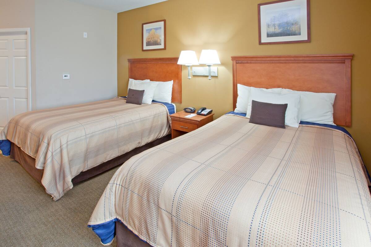 Quarto de hotel com duas camas, uma mesinha de cabeceira e uma luminária fixada na parede.