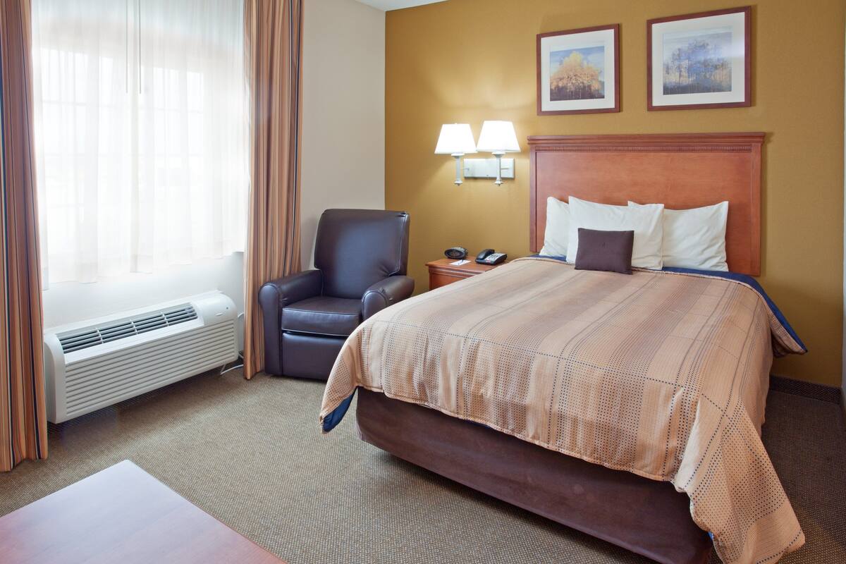 Quarto de hotel com uma cama grande, uma cadeira, uma escrivaninha e uma janela com cortinas.