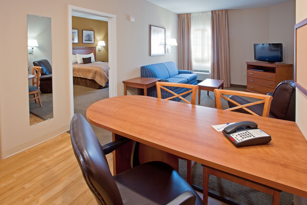 Habitación de hotel con una mesa de comedor de madera, un sofá azul, una cama con cabecera marrón, un televisor sobre un mueble de madera y un teléfono sobre la mesa.