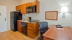 A compact kitchen with a refrigerator, microwave, and coffee maker, a wooden dining table with a chair, and a wall-mounted lamp.