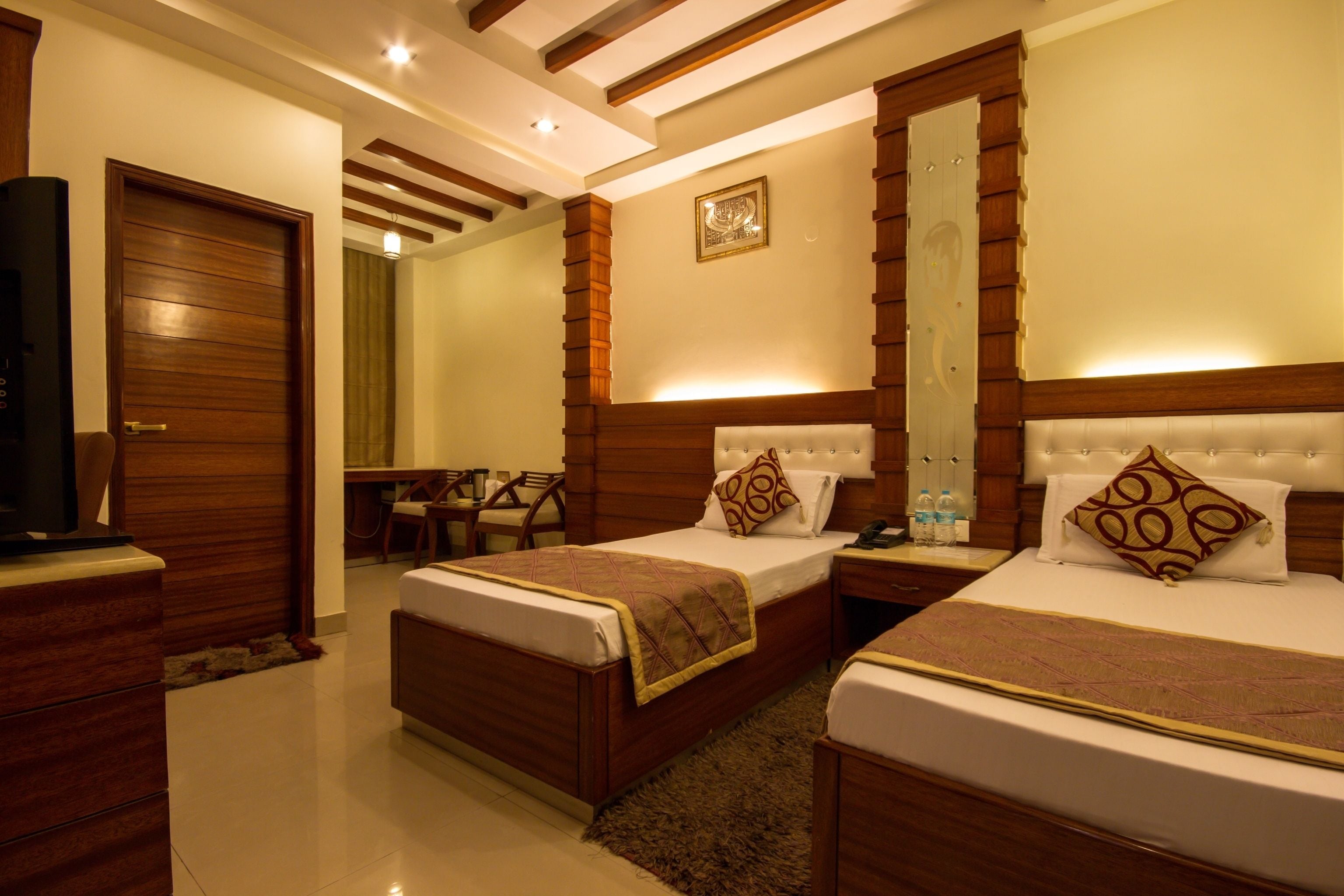 A hotel room with two beds, wooden headboards, a bedside table with a lamp, a mirror, and a small seating area with a chair and table.