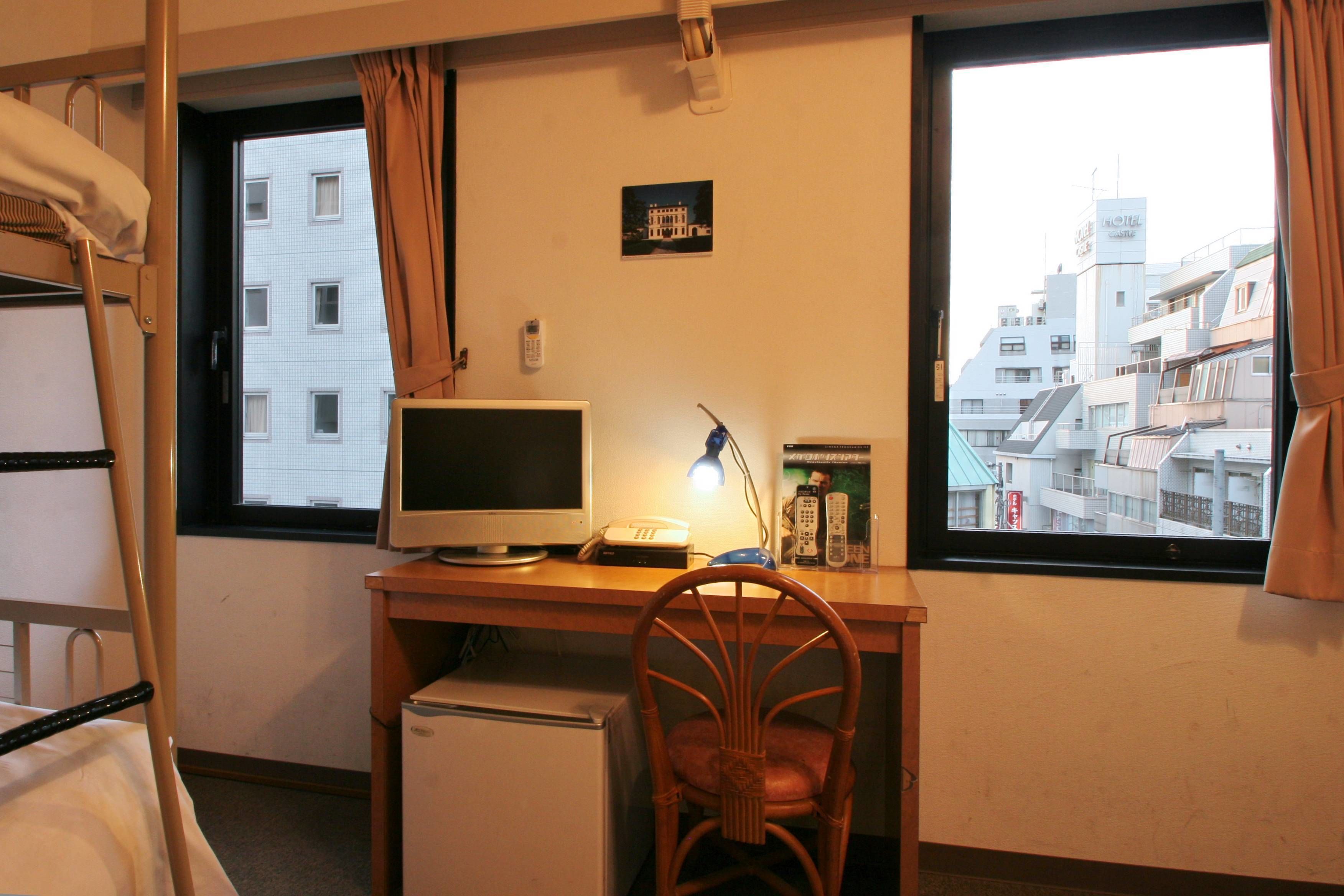 Ein Hotelzimmer mit einem Bett, einem Schreibtisch mit Computer, einem Sessel, einem kleinen Kühlschrank und einem Fenster mit Blick auf Gebäude.
