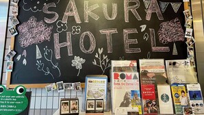A hotel reception area with a chalkboard sign reading "SAKURA HOTEL," various travel brochures, and a notice about umbrella storage. - Sakura Hotel Ikebukuro - Hostel (Tokyo)