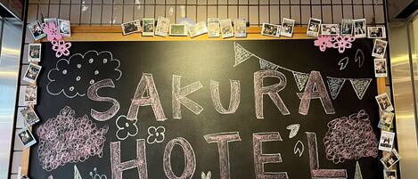 A hotel reception area with a chalkboard sign reading "SAKURA HOTEL," various travel brochures, and a notice about umbrella storage.