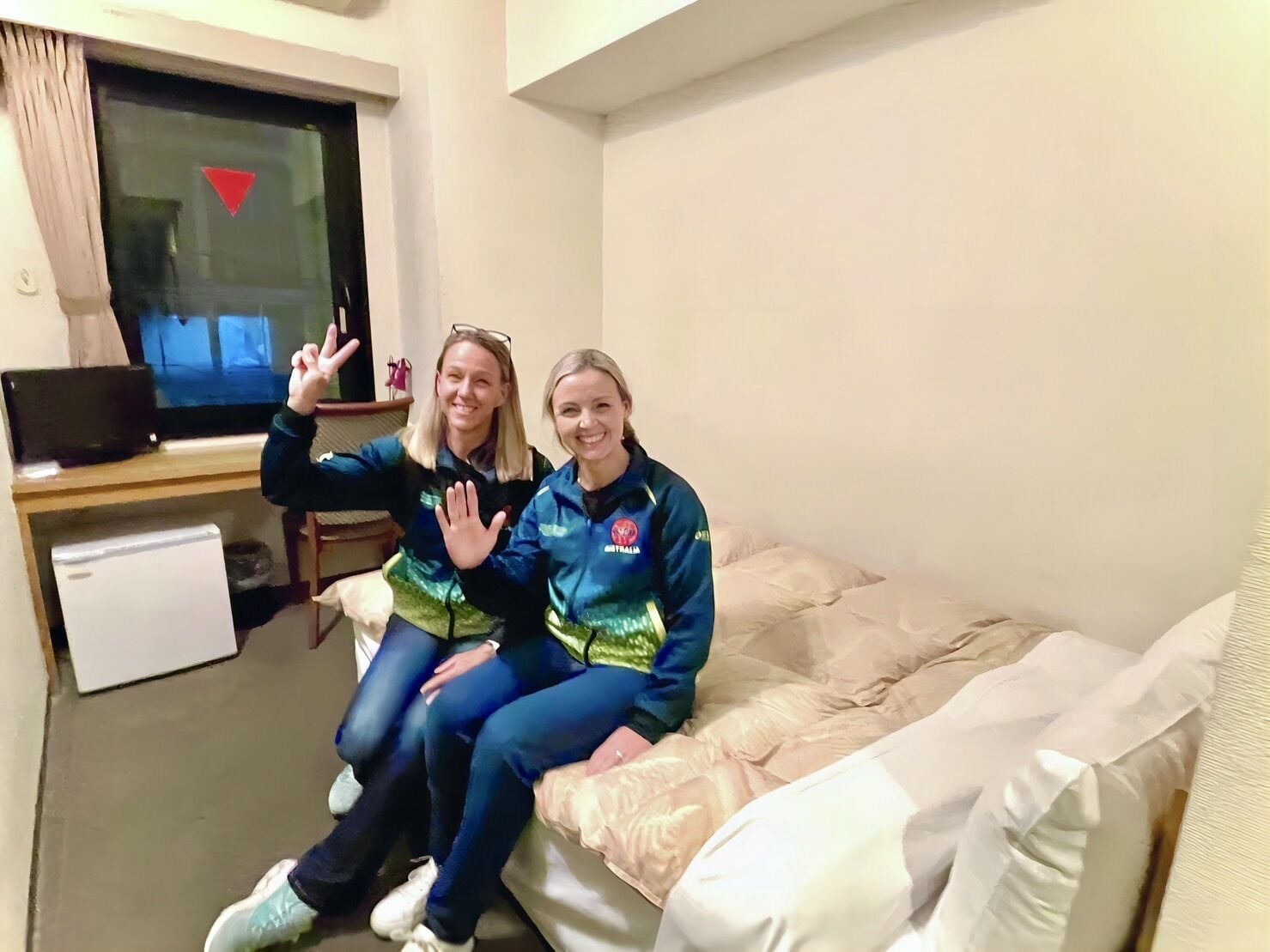 Two people sitting on a bed in a room with a television, a small fridge, and a window with a red triangle symbol.