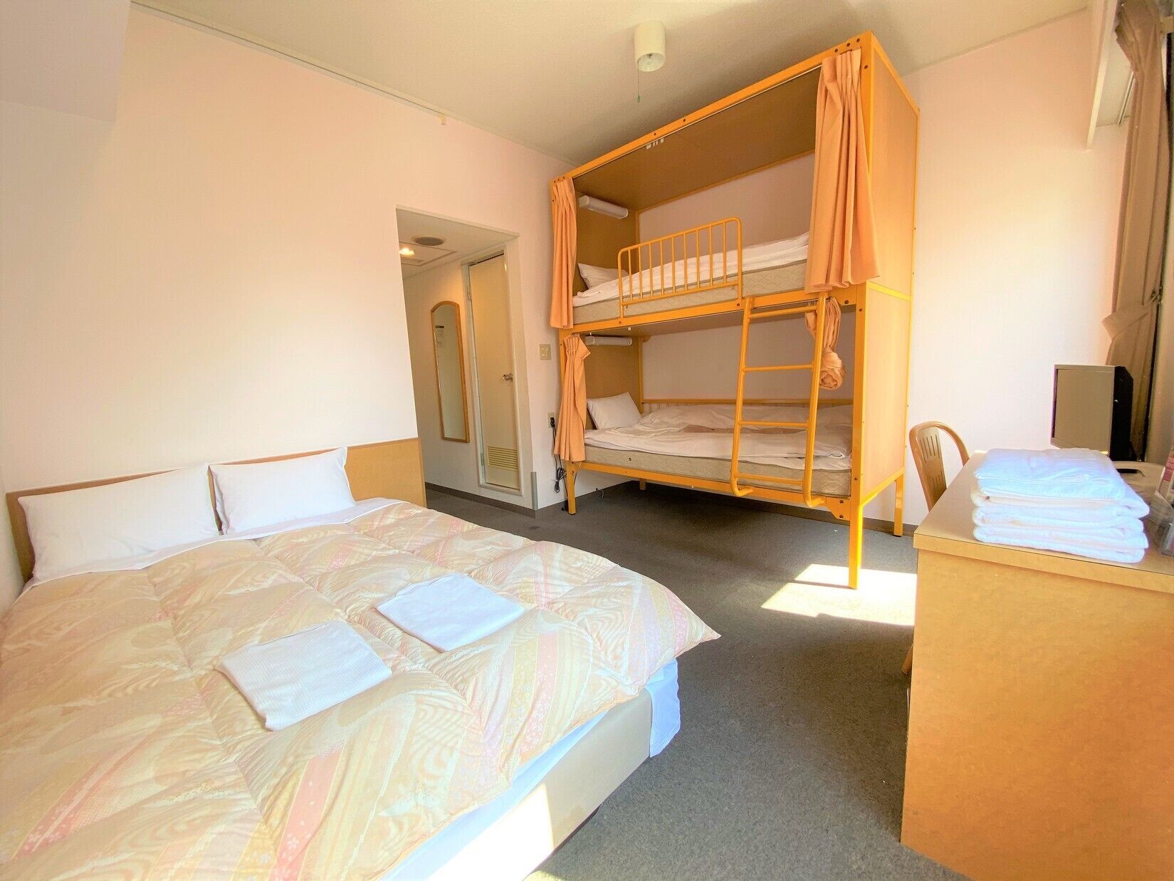 A hotel room with a single bed, a bunk bed, a desk with a chair, and a shower area.