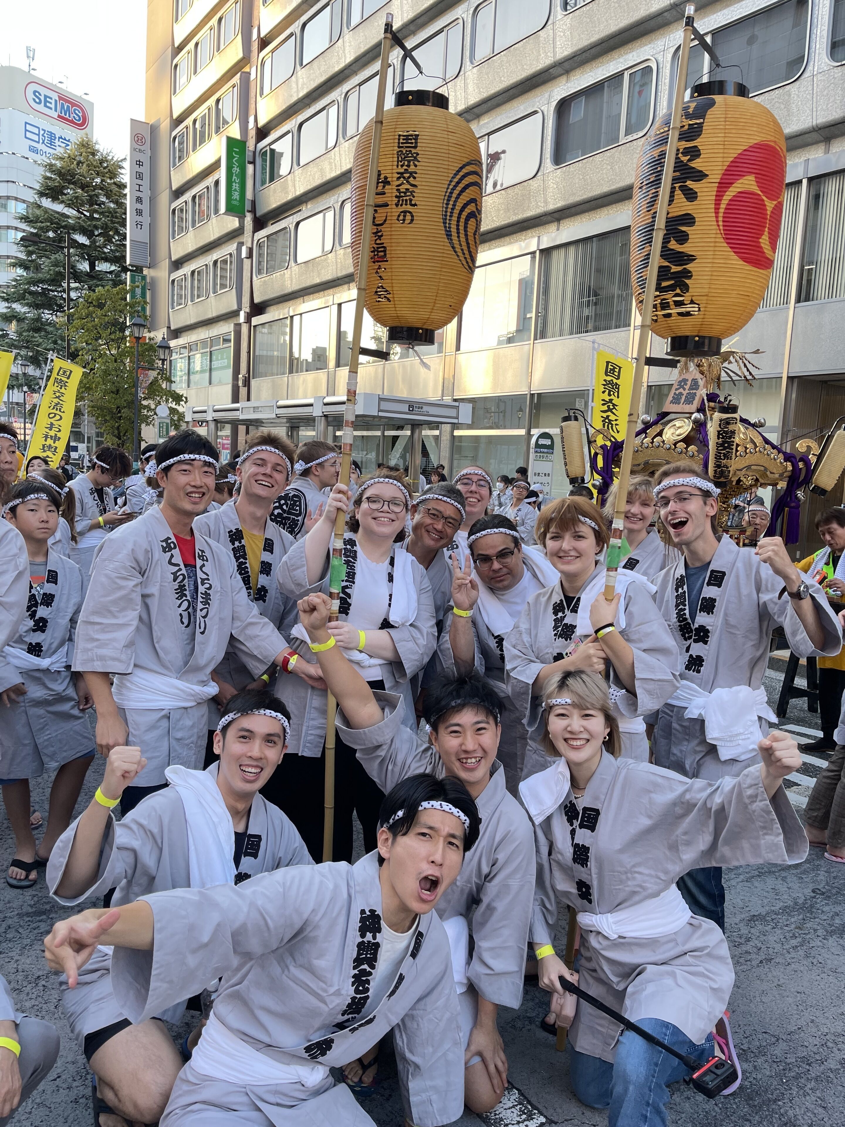 A group of people in traditional attire, some holding banners with Japanese text, gathered in an urban setting with modern buildings in the background.