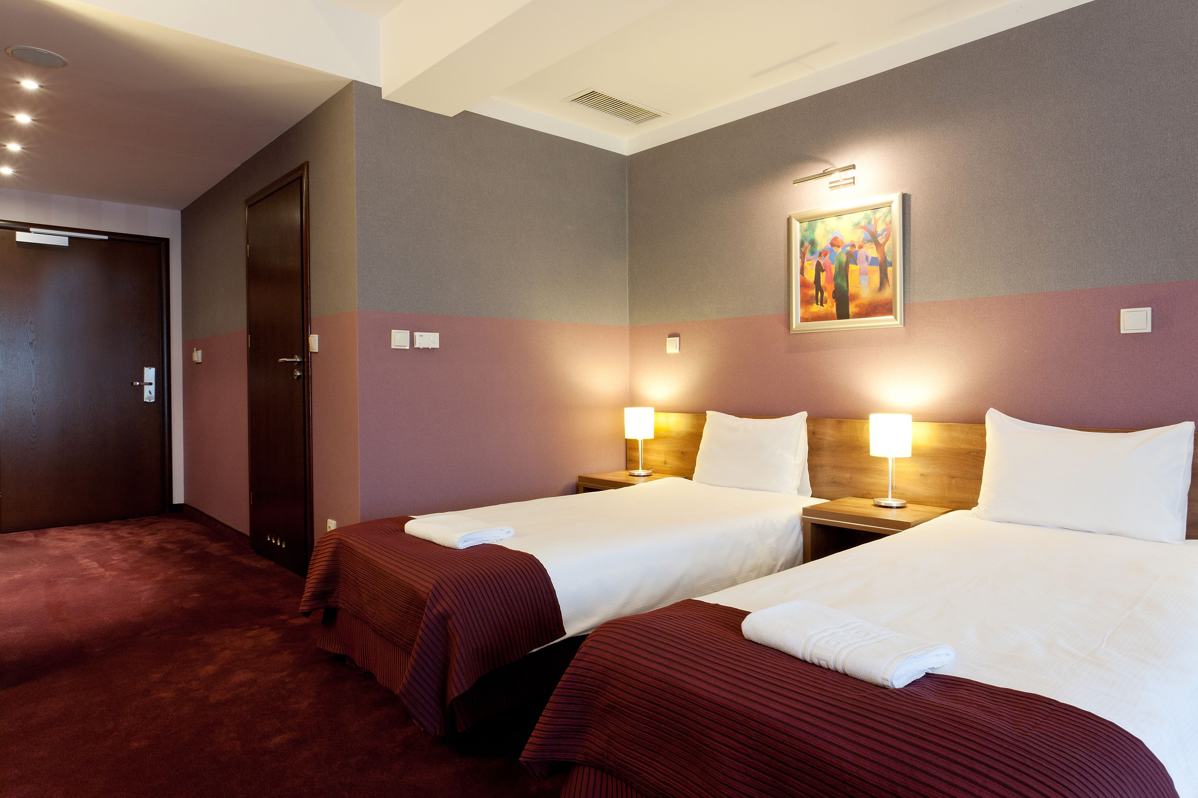 A hotel room with two beds, a door, and a painting on the wall.