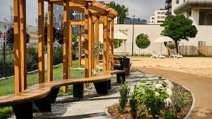 Een houten pergola met banken, een verhard pad en een aangelegde tuin met bloemen en een boom.