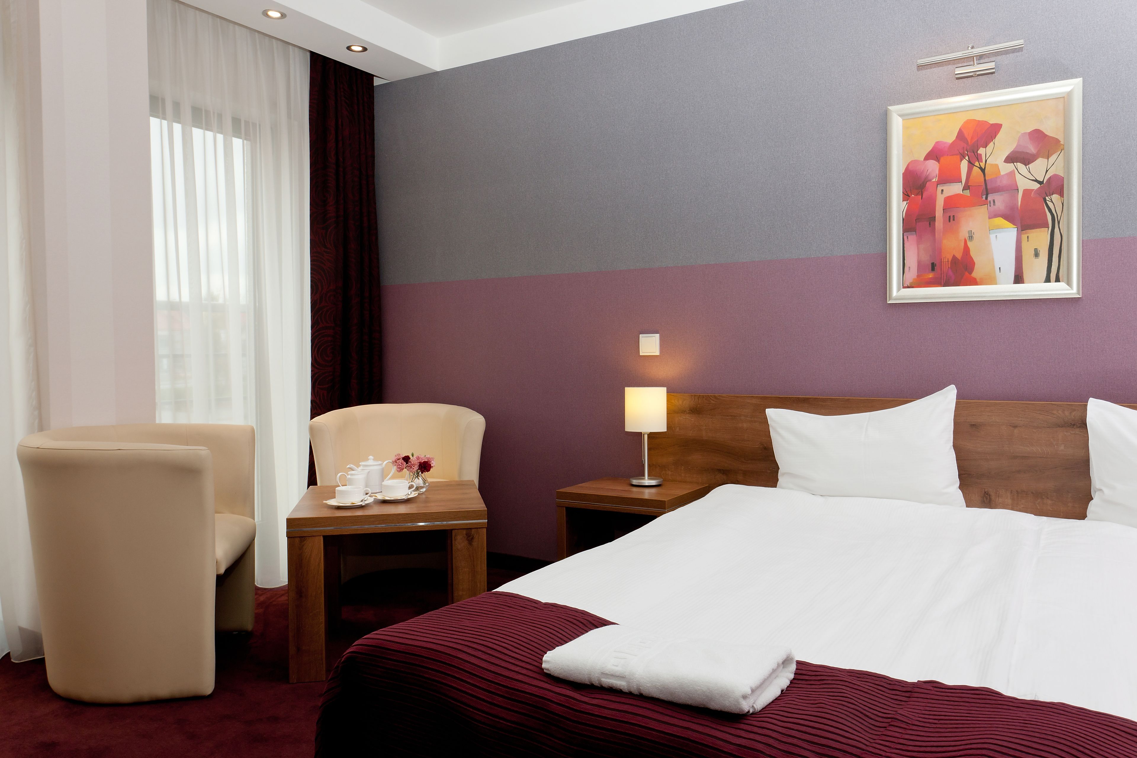 Una habitación de hotel con una cama, una silla, una mesita de noche con lámpara y un cuadro en la pared.