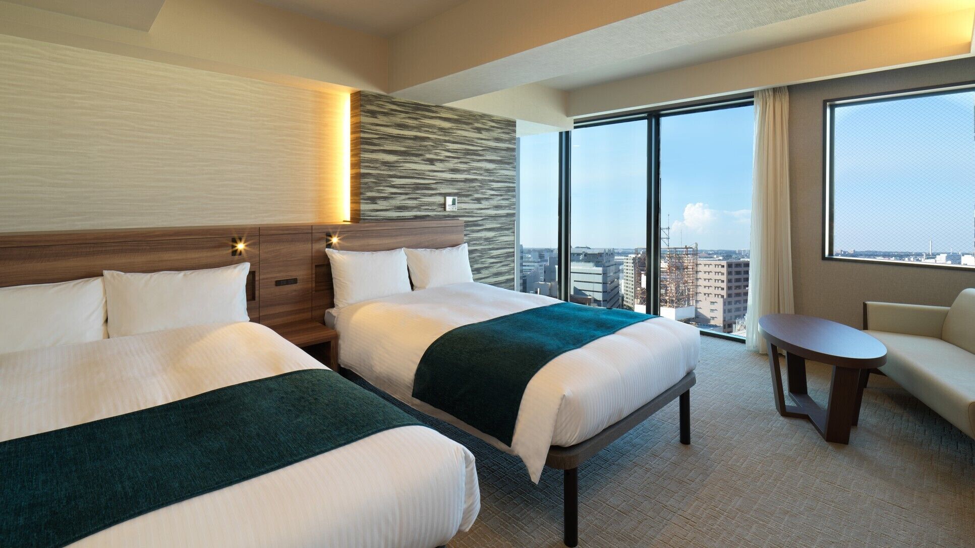 A hotel room with two beds, a sofa, a small table, and a large window offering a city view.