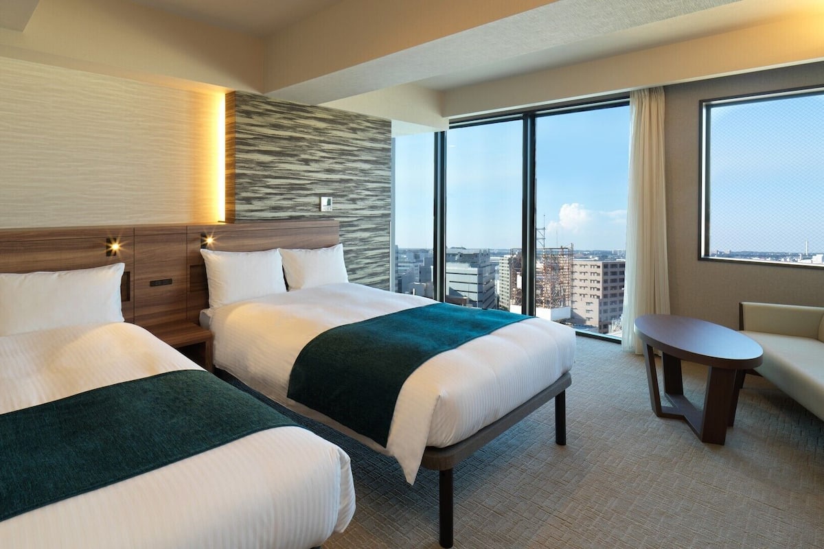A hotel room with two beds, a sofa, a small table, and a large window offering a city view.