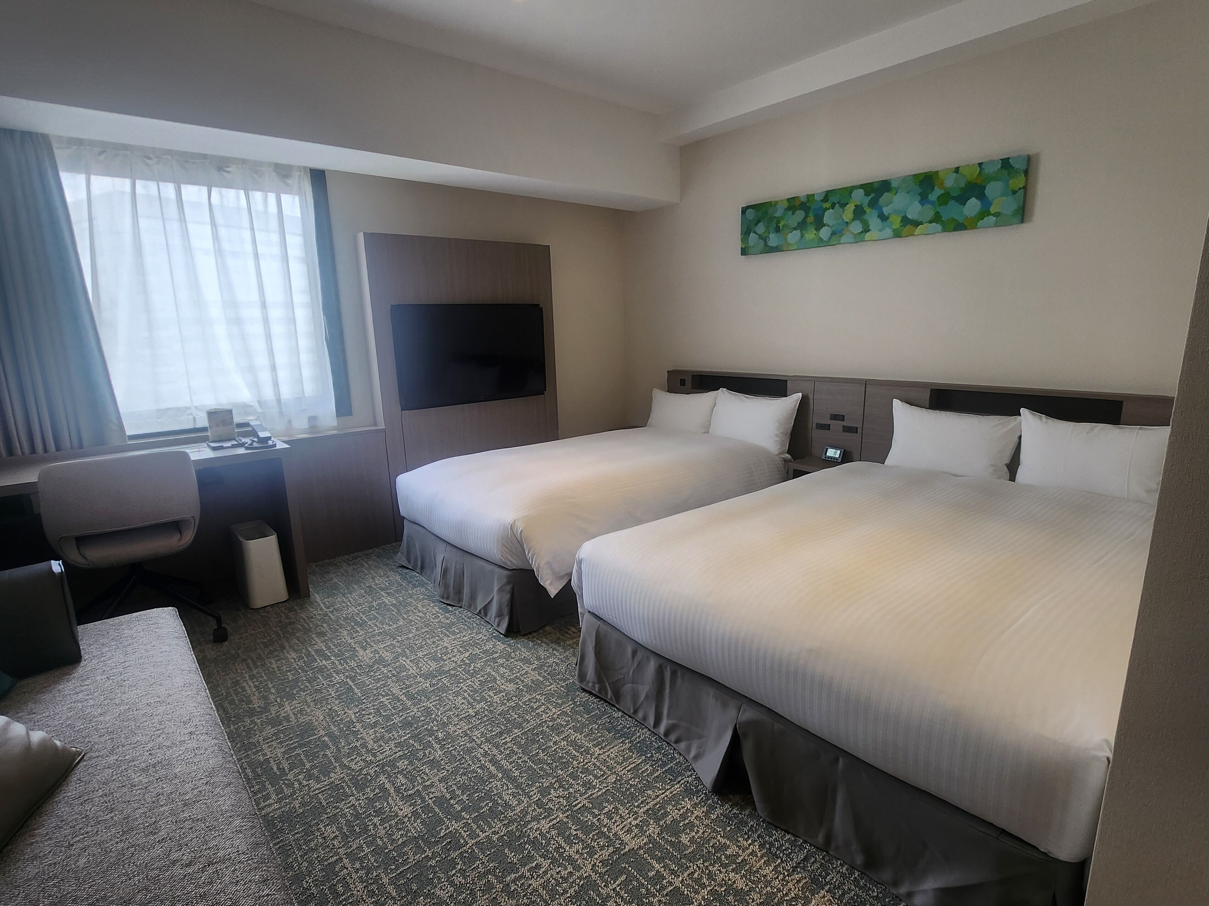 A hotel room with two beds, a desk, a chair, a TV, and a painting on the wall.